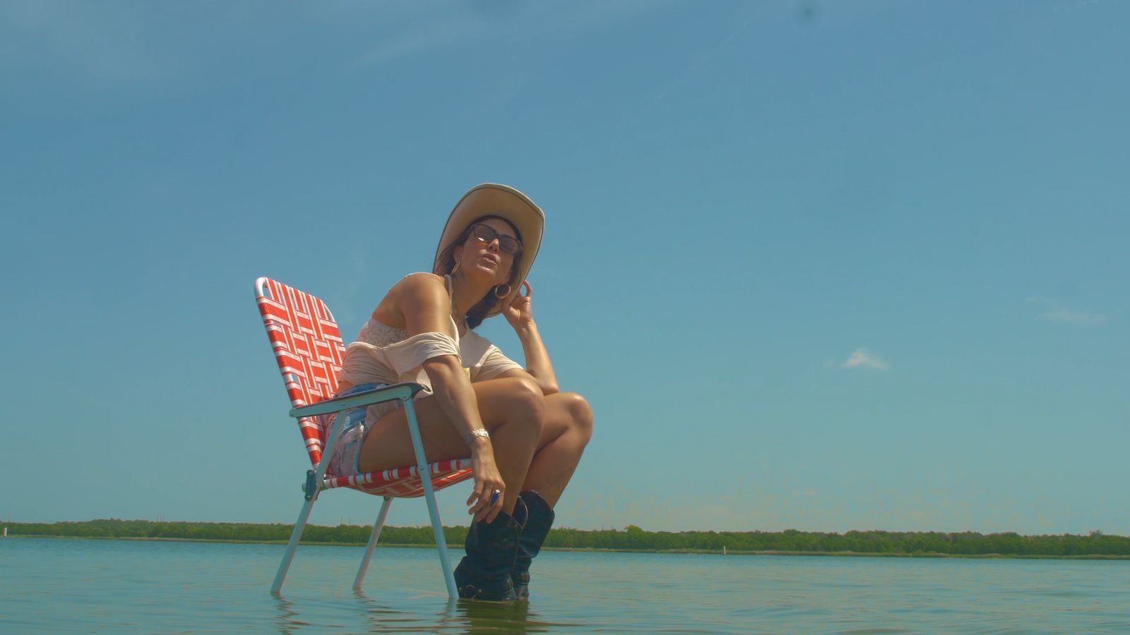 JE55IE sits in a red and white striped lawn chair standing in shallow lake water, wearing a cream cowboy hat, white tee, denim cutoffs and black boots, against a wide open blue sky.